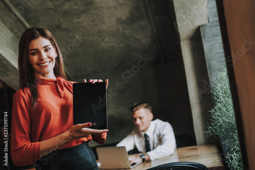 Concept of successful team work. Waist up portrait of smiling business lady showing screen of tablet computer while her partner working on laptop
