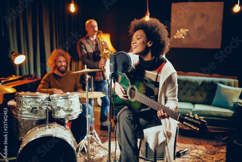 Mixed race woman singing and playing guitar while sitting on chair with legs crossed. In background drummer, saxophonist and bass guitarist.