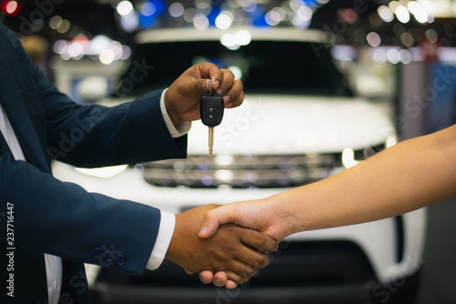 Handshake of two businessmen when selling a car in a motor show, auto business, car sale, deal, gesture and people concept - close up of dealer giving key to new owner and shaking hands in showroom.