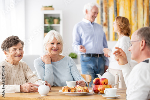 Senior people visiting old friends at home sitting together at wooden table chatting, drinking tea and eating cake