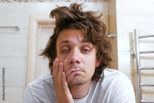 Sleepy shaggy young man looks at the mirror in bathroom in the morning.