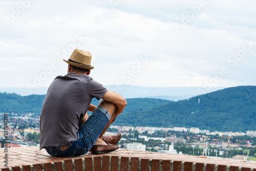 A young guy in comfortable moccasins sits on the edge of a tall building and looks at the mountains and the city of Graz