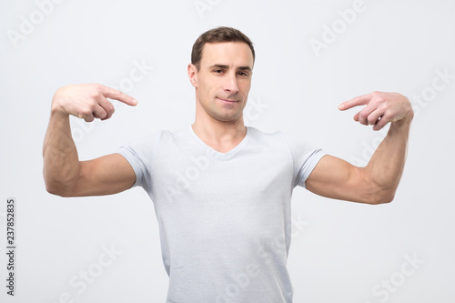 Italian man in white t-shirt, standing, pointing himself and looking with serious egoist face.