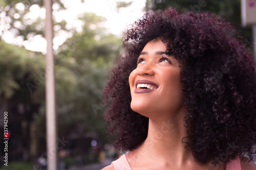 Portrait of woman looking up and smiling. Concept of imagination, hope, planning. Outdoors. She is black, on her early twenties, Afro style frizzy hair.