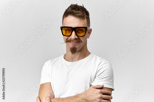 Cheerful attractive young unshaven male with stylish hairdo, goatee beard and handlebar mustache being in good mood, crossing arms on his chest and smiling confidently, wearing trendy spectacles