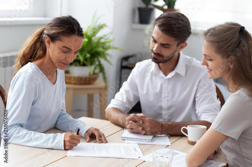 Mixed race client hold pen put signature at official paper make buy or sell deal sitting with partners during meeting. After successful job interview millennial woman signing employment agreement