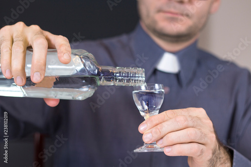 Ksiądz w szarej koszuli i koloratce nalewa wódkę z butelki do kieliszka.