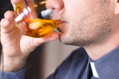 Ksiądz w koloratce i szarej koszuli pije whisky z lodem ze szklanki.
