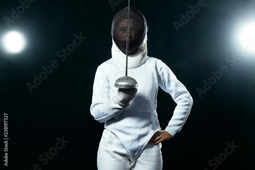 Young fencer athlete wearing fencing costume holding the sword and mask. Isolated on black background with lights