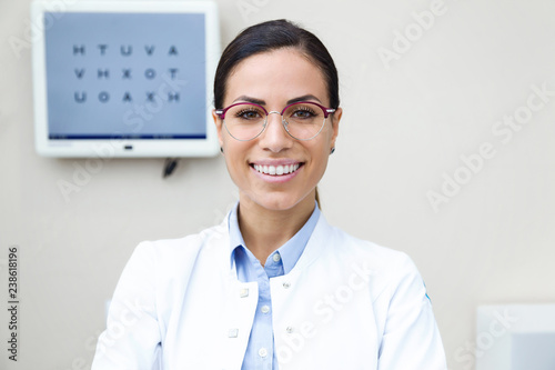 Pretty young woman ophthalmologist smiling while looking at camera in the consultation.