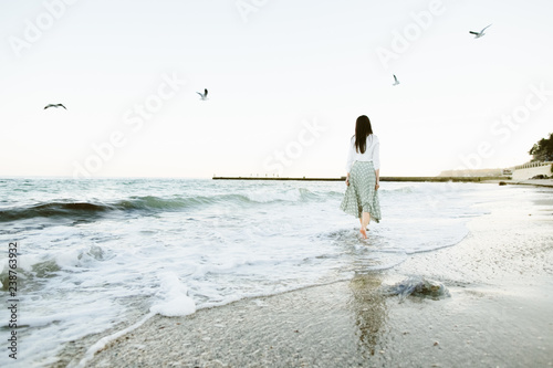 Marine female portrait. Attractive woman in green skirt walks along the shore before the sea while gulls fly over her