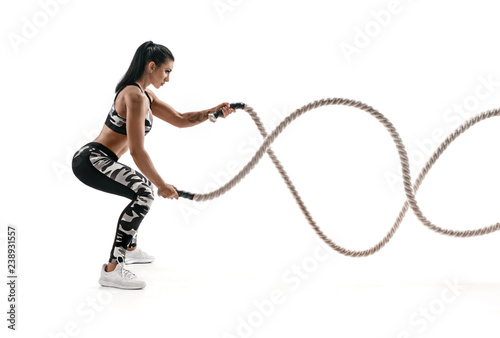 Strong muscular woman working out with battle ropes. Photo of attractive woman in fashionable sportswear isolated on white background. Strength and motivation. Side view.