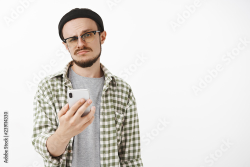 Indifferent male young snob in glasses and beanie with beard holding smartphone smirking and staring at camera careless and unimpressed receiving offer via messages and being dissatisfied