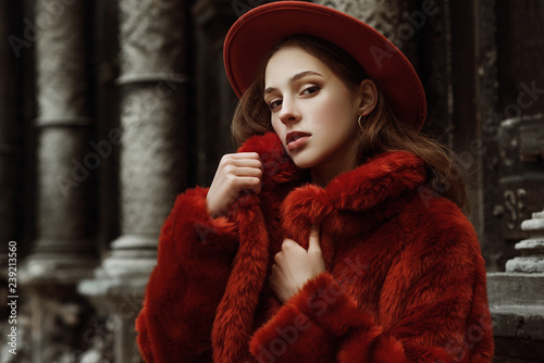 Outdoor close up fashion portrait of young beautiful confident woman wearing trendy orange faux fur coat, hat, silvery hoop earrings, posing in street of european city. Copy, empty space for text 