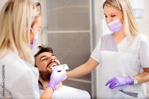 Two young female dentists working in dental clinic. Whitening male patient's teeth and using tone chart to mach color. Health care and medicine concept.