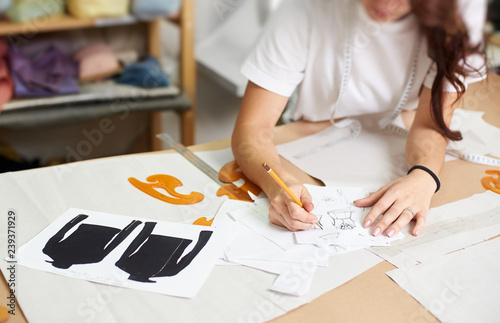 Female designer drawing pencil sketches of clothing sitting at big table with flat paper patterns, measuring ruler and curves on blurred background of tailor design studio interior.