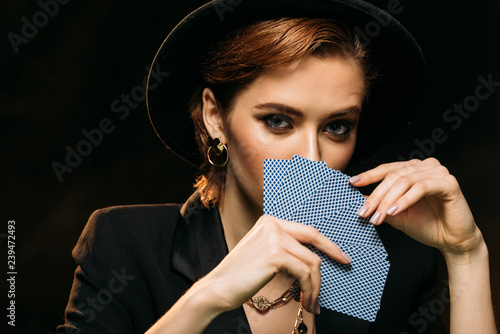 attractive girl in jacket and hat covering face with poker cards isolated on black, looking at camera