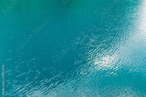 Shiny texture of azure surface of mountain lake. Minimalist background with reflection of mountains and green trees in clear water in sunny day. Copy space.