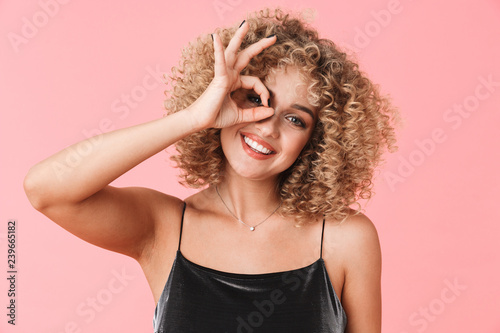 Portrait of amusing curly woman 20s wearing dress smiling and showing ok sign while standing, isolated over pink background