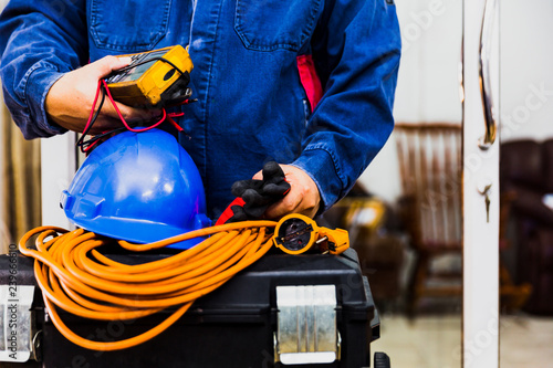 Electrian engineer holding multimeter and tools in hand, standing behind the heavy duty tool box, image including power cord, Blue hard hat (helmet) and gloves.