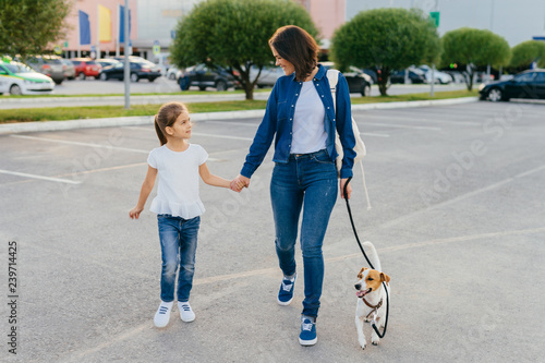 Photo of happy daughter and mother have outdoor stroll against city background, walk their dog, keep hands together, spend weekend together, enjoy spare time. Family, animals and leisure concept