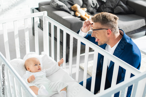 happy father in formal wear showing tongue to infant daughter in baby crib