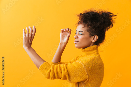Portrait of beautiful african american woman with afro hairstyle looking through invisible spyglass, isolated over yellow background