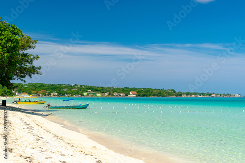 Sunny day along the Seven Mile Beach in tropical Negril, Jamaica. Tour boats await passengers and caucasian tourists in the water at a distance.