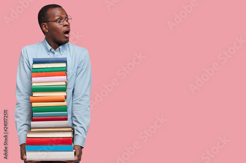 Studio shot of stupefied dark skinned black male professor, holds many books neatly arranged, prepares for seminar or conducting lecture, dressed formally, isolated on pink wall with copy space