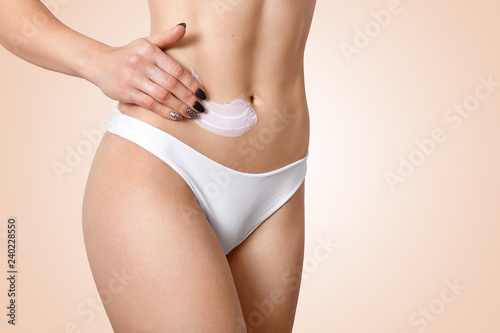Young fit woman applies cosmetic cream for cellulite or lotion on belly, wears white panties, cares of skin, isolated over beige background, avoids stretching. Cosmetology and medicine concept
