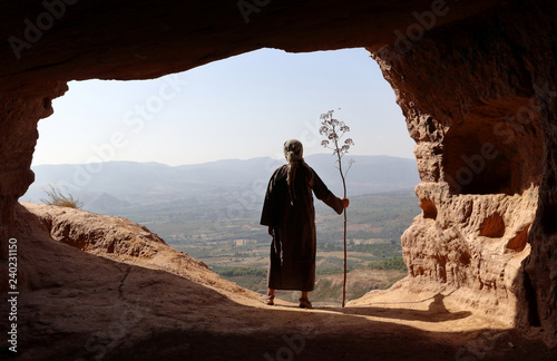 HERMIT MAN LOOKING TO THE VALLEY FROM A HIGH CAVE WITH A BRANCH IN THE HAND IN SUMMER 