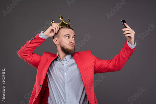 Young bearded handsome man in gold crown taking selfie looking at smartphone