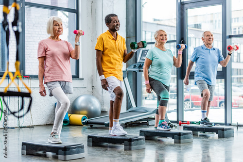 smiling senior multicultural sportspeople synchronous exercising with dumbbells on step platforms at gym