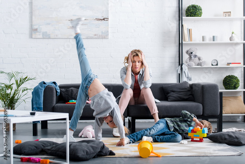 tired mother sitting on couch and looking at naughty kids playing at home