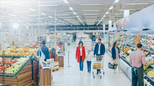 At the Supermarket: Happy Family of Three, Holding Hands, Walks Through Fresh Produce Section of the Store. Father, Mother and Daughter Having Fun Time Shopping. High Angle Panoramic Shot.