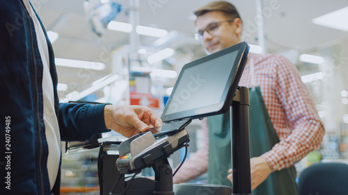 At the Supermarket: Checkout Counter Customer Pays with Smartphone for His Food Items. Big Shopping Mall with Friendly Cashier, Small Lines and Modern Wireless NFC Paying Terminal System.