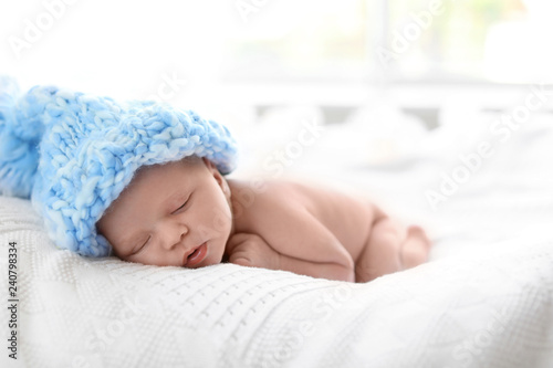 Adorable newborn baby in large knitted hat lying on bed against light background