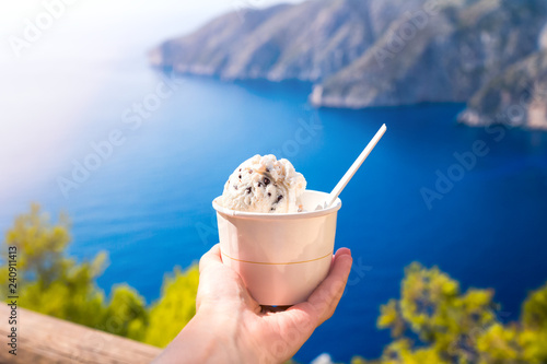 Hand holding ice cream over blue sea and white cliff from above, the view of a bay in Greek island, travel destination