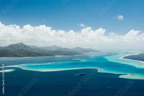 Aerial view on lagoon of Raiatea island in French Polynesia with blue and turquoise water, barrier reef, blue sky, hills with tropical forest and white clouds