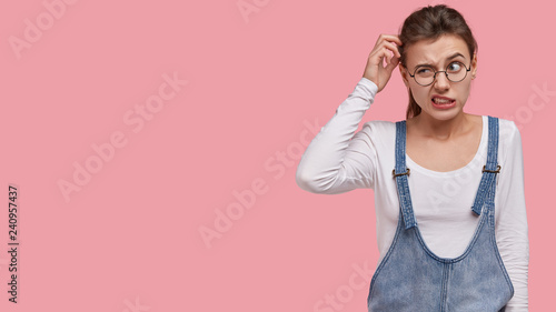 Photo of puzzled woman scratches head in bewilderment, thinks about finding right solution, wears trendy sarafan, round spectacles, models over pink background with free space for information