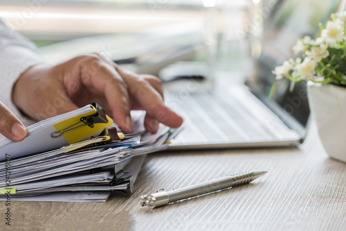 Businessman hands holding pen for working in Stacks of paper files searching information business report papers and piles of unfinished documents achieves on laptop computer desk in modern office