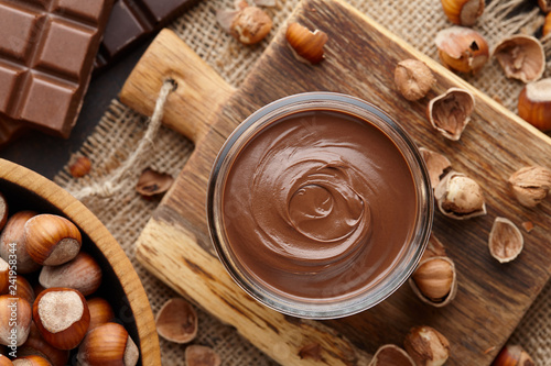 Chocolate spread or nougat cream with hazelnuts in glass jar, bowl of nuts and chocolate bars on brown countertop