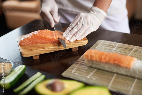 Close-up view of process of preparing delicious rolling sushi in restaurant. Female hands in disposable gloves slicing salmon.