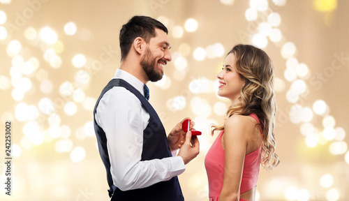 love, couple, proposal and people concept - happy man giving diamond engagement ring in little red box to woman over festive lights background