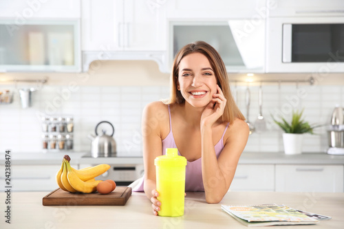 Young woman holding bottle of protein shake at table with ingredients in kitchen