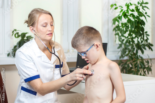 Lekarka pediatra w białym kitlu bada stetoskopem chłopca w niebieskich okularach.