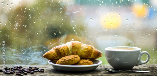 Fresh brewed coffee in white cup or mug on windowsill. Coffee drink with croissant dessert. Enjoying coffee on rainy day. Coffee time on rainy day. Wet glass window and cup of hot caffeine beverage