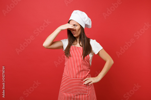 Housewife female chef cook or baker in striped apron, white t-shirt, toque chefs hat isolated on red wall background. Housekeeper woman close nose with hand because smell. Mock up copy space concept.