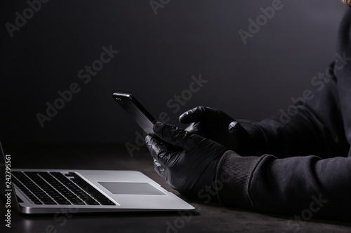 Professional hacker with laptop and mobile phone sitting at table on dark background