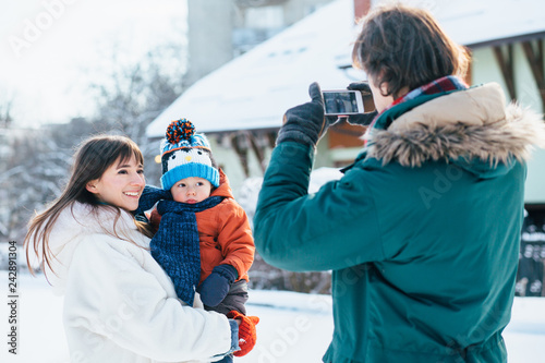 Father takes pictures on phone of happy mother and little baby toddler son in her arms in snowy winter day outdoor.
